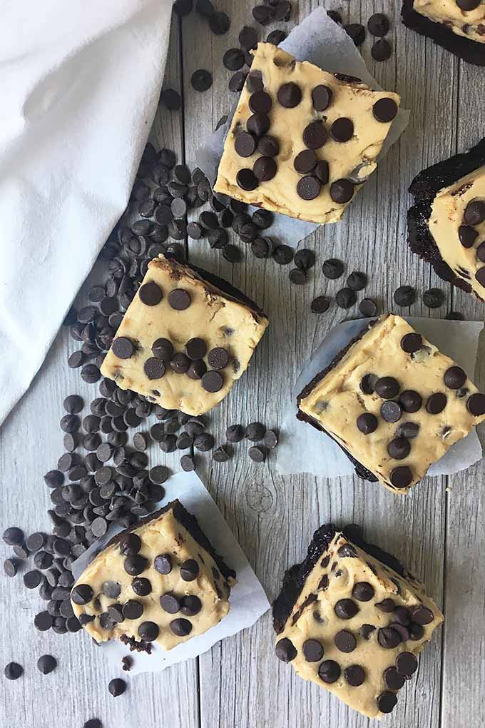Vertical top-down image of cookie dough bars on top of pieces of parchment paper surrounded by chocolate chips on a white wooden surface.