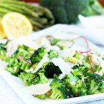 A white, square plate of sautéed asparagus and broccoli with thinly sliced radishes and parmesan cheese, with whole vegetables and a cut lemon in soft focus in the background.