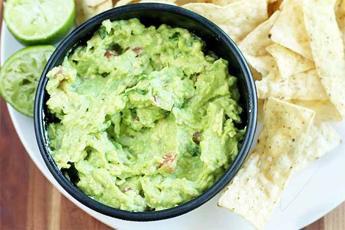 Top-down shot of a black bowl of green guacamole with tortilla chips and a lime cut in half with one half that has been juiced already, on a brown wood background.