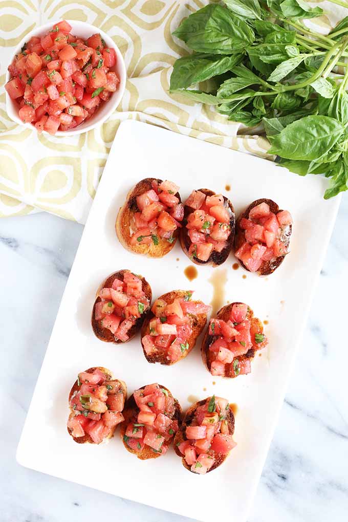 Top-down view of nine pieces of toasted baguette rounds topped with chopped tomatoes and basil, and drizzled with balsamic vinegar, on a rectangular, white, ceramic serving dish, on a marble surface topped partially with a beige and white patterned cloth, will a small dish of bruschetta topping and a few sprigs of green fresh basil.