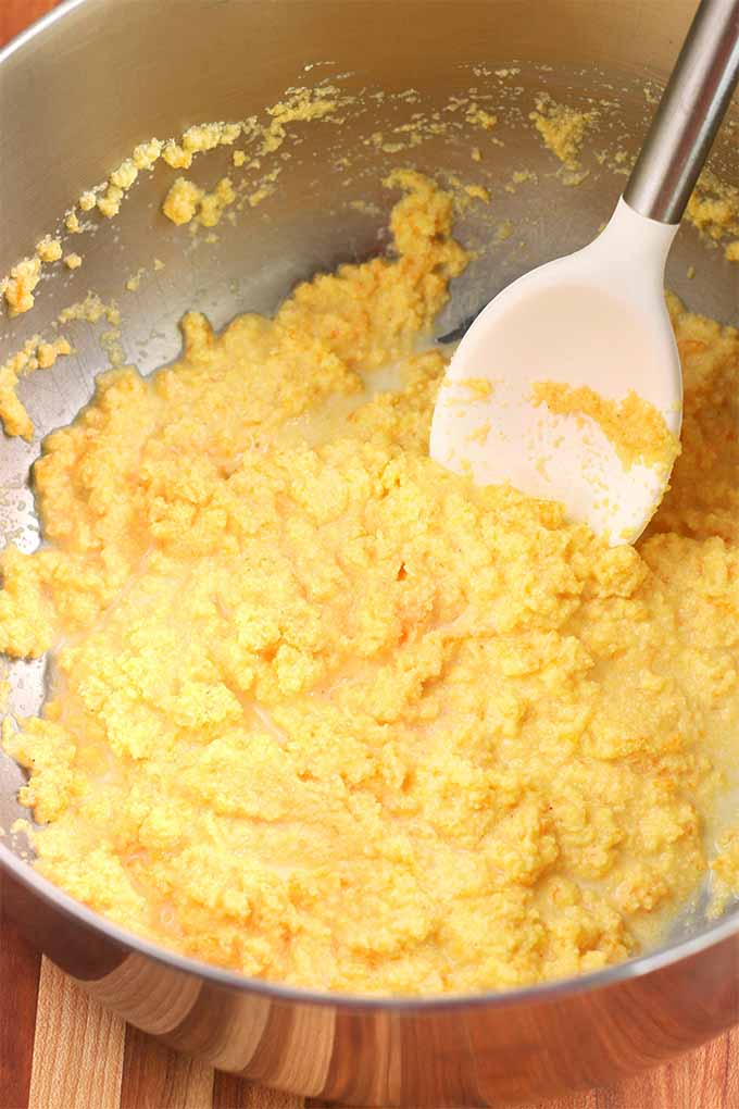 Top-down closely cropped shot of a yellow cornmeal batter mixture in a stainless steel bowl with a white rubber spatula.