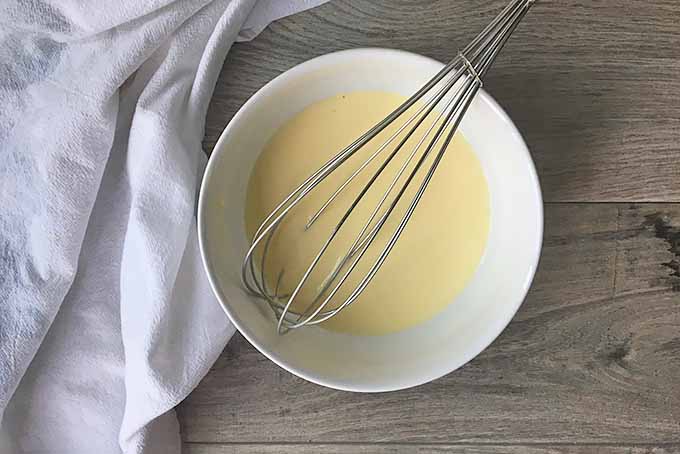 Horizontal image of a whisk in a yellow mixture in a white bowl.