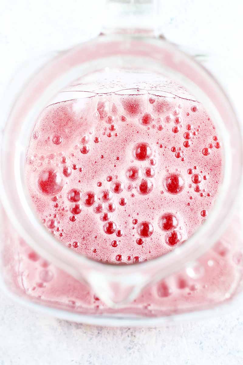 Top-down vertical image of a bubbly pink liquid in a clear g.ass pitcher, on a blue and white speckled background.