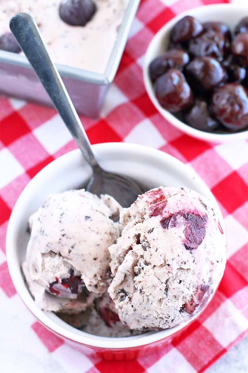 Top-down shot of a scoop of ice cream in a ramekin with a spoon, beside a loaf pan of more of the dessert and a small white bowl of black cherries behind it, on a white and red checkered cloth.