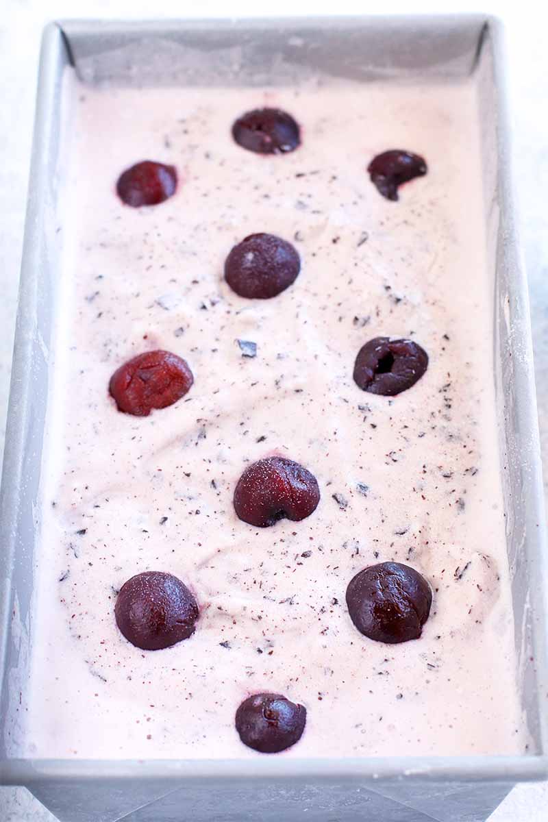 Top-down view of a metal loaf pan filled with pink homemade ice cream, speckled with chocolate and topped with glazed black cherry halves arranged in a diamond pattern.