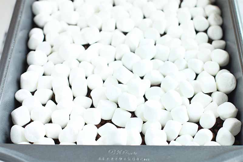 White miniature marshmallows are scattered on top of a layer of chocolate in a gray metal baking pan.