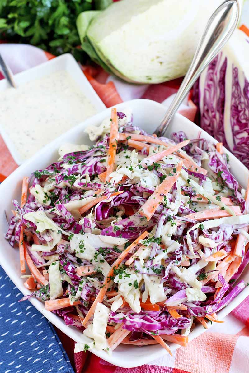 Oblique vertical view of a square bowl with rounded corners, filled with homemade cabbage slaw, with vegetables and a dish of dressing in the background, and a folded blue cloth napkin in the foreground.