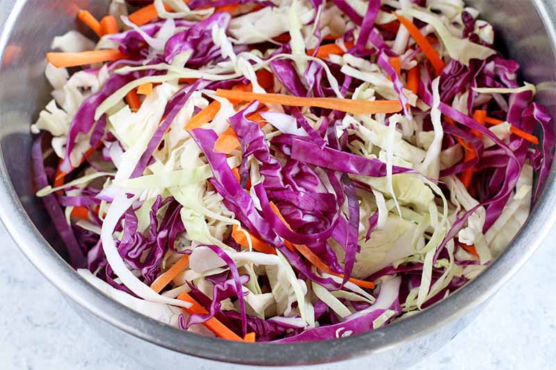 A large stainless steel mixing bowl of shredded purple and green cabbage and carrots.