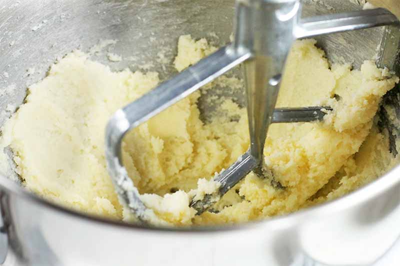 A pale yellow mixture of creamed butter and sugar in a stainless steel stand mixer bowl with a matching silver paddle attachment.