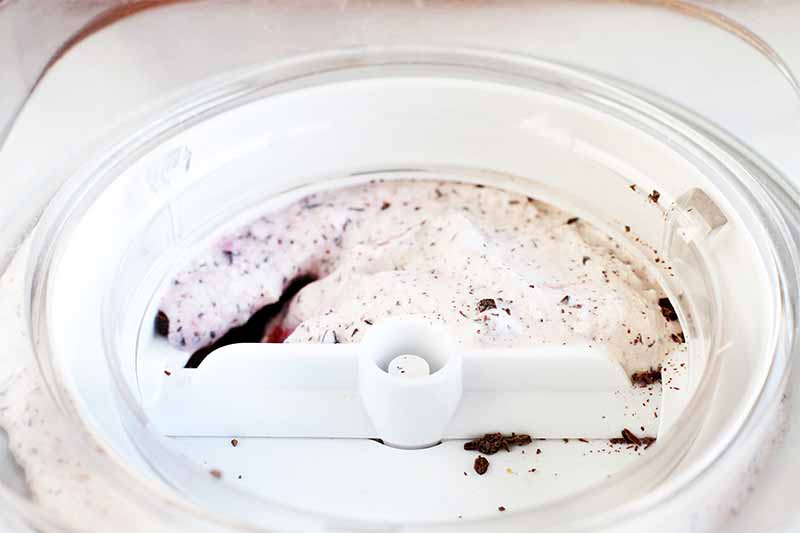 A white plastic dasher rests at the center of a white and gray freezer container of pink cherry and chocolate ice cream.