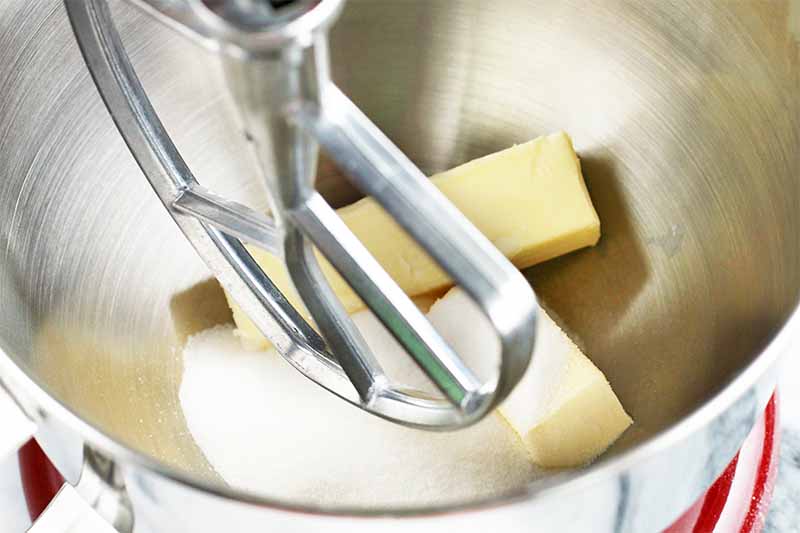 Two sticks of butter and a small pile of sugar at the bottom of a stainless steel stand mixer bowl with a matching paddle attachment.
