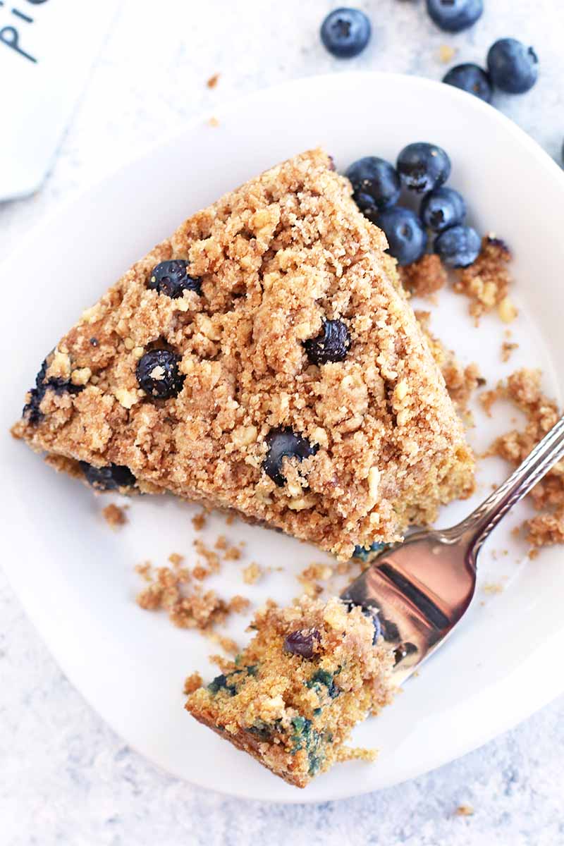 A slice of blueberry crumb cake on a plate, with a fork holding a bite up towards the camera, with scattered berries on a white background.