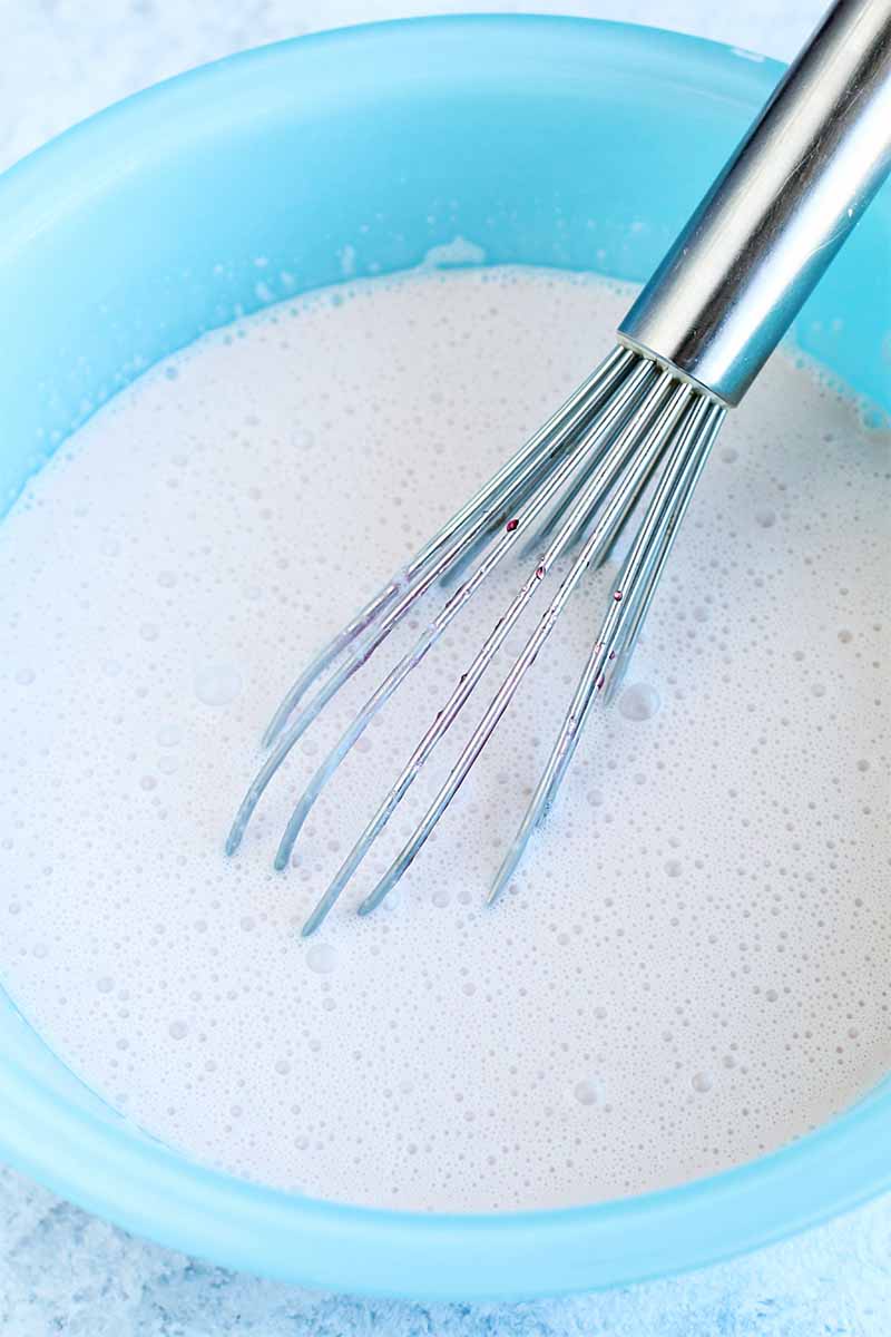 Top-down shot of a light blue glass bowl of a white cream mixture, with a silver-colored wire whisk.