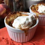 Horizontal image of cobbler topped with whipped cream on an orange towel with peaches and spices in the background.