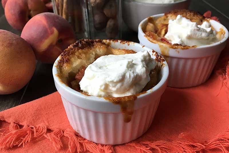 Horizontal image of cobbler topped with whipped cream on an orange towel with peaches and spices in the background.