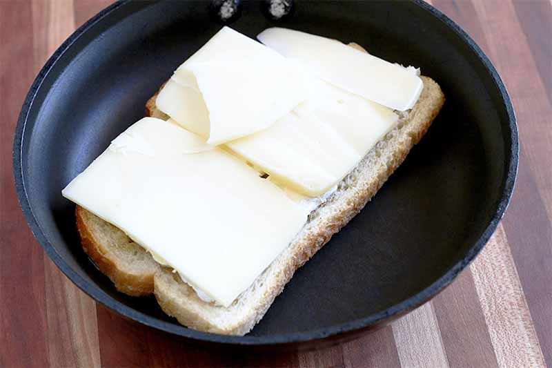 Thinly sliced gruyere cheese is arranged on a piece of sourdough bread in a nonstick frying pan.