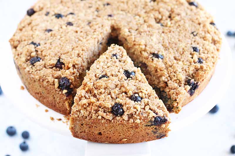 A round blueberry crumb cake with a triangular slice taken out with a cake server, on a white background with scattered berries.
