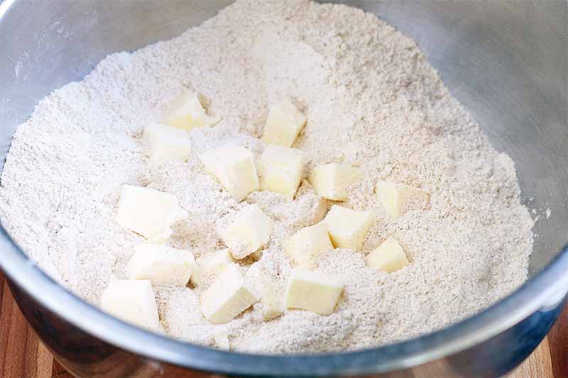 Cubed butter and flour in a stainless steel mixing bowl, on a brown wood surface.