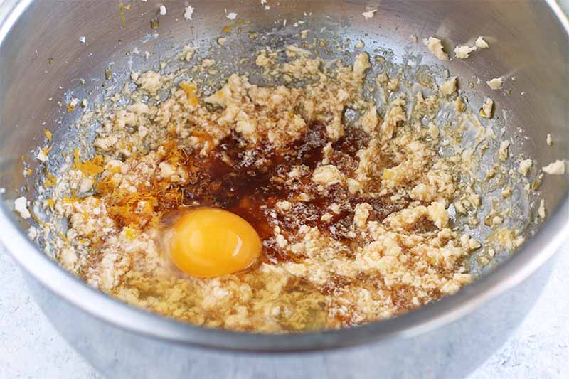 A raw egg, maple syrup, and vanilla in a creamed butter and sugar mixture in a stainless steel mixing bowl.