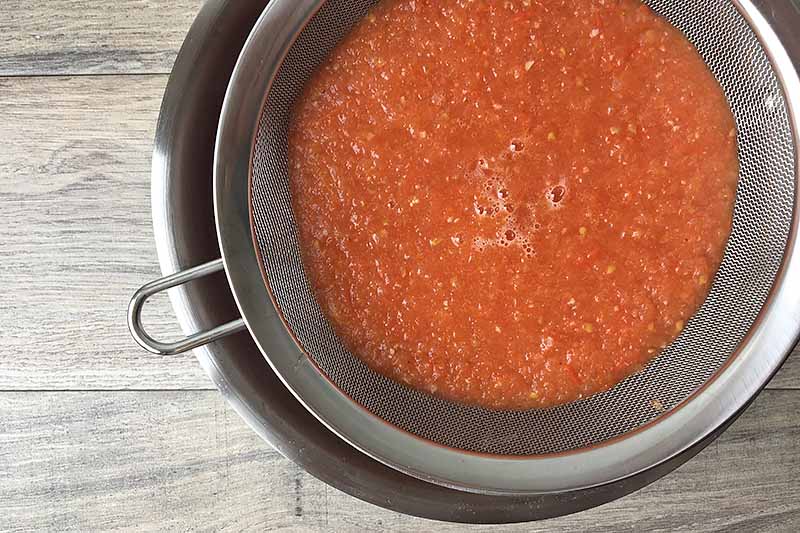 Horizontal image of a lumpy red sauce in a strainer over a bowl.