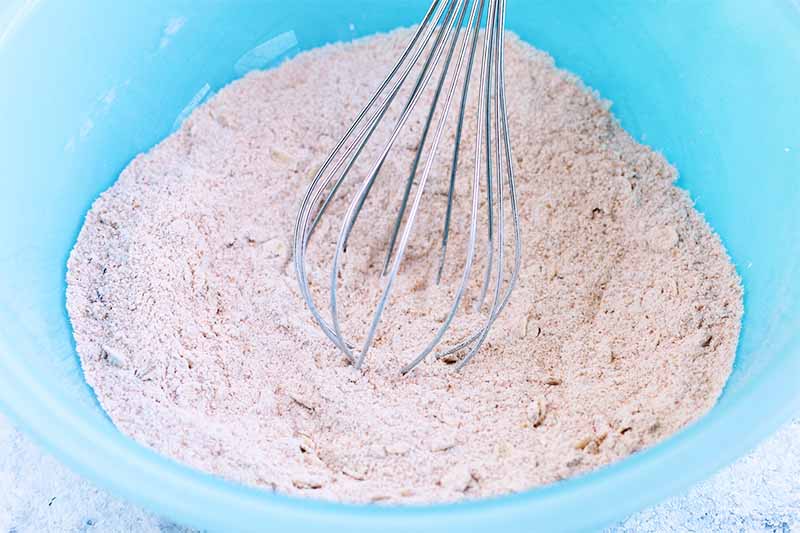 A silver wire whisk stirs a whole wheat flour mixture in a light blue bowl.