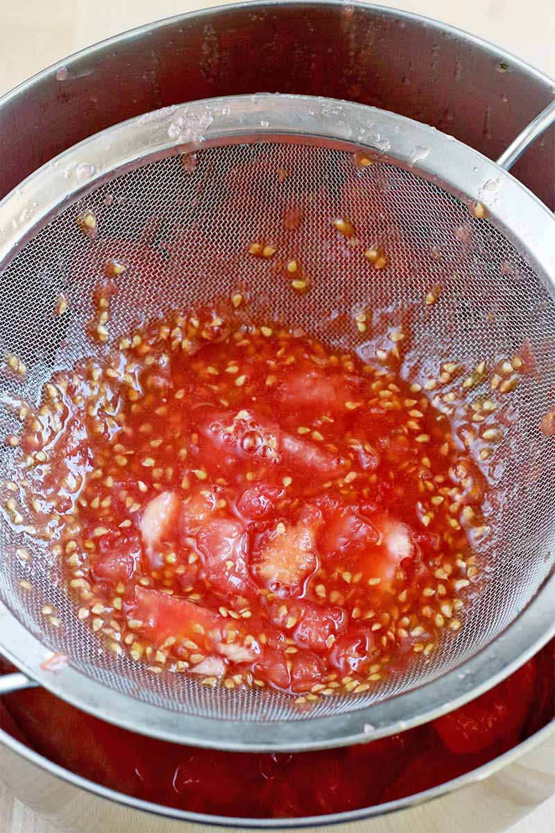 Tomato seeds and juice in a mesh strainer, resting on top of a stainless steel bowl.