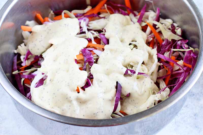 A large stainless steel bowl of shredded cabbage and carrots, drizzled with a white mayonnaise sauce.