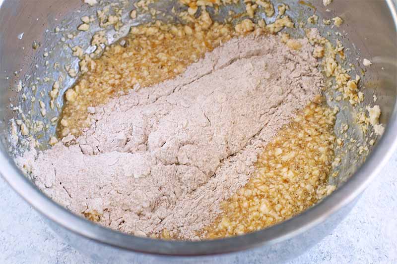 Dry whole wheat flour is sprinkled on top of a creamed butter wet mixture in a large stainless steel mixing bowl.