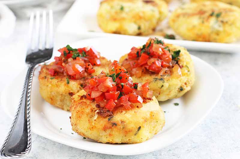 Three potato fritters topped with red bell pepper relish on a white plate with a fork, with more in the background on a serving platter.
