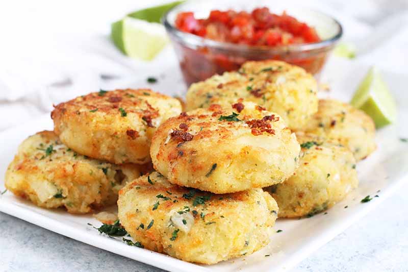 Golden brown mashed potato fritters stacked on a white rectangular serving platter with lime wedges and a small glass dish of red pepper relish.