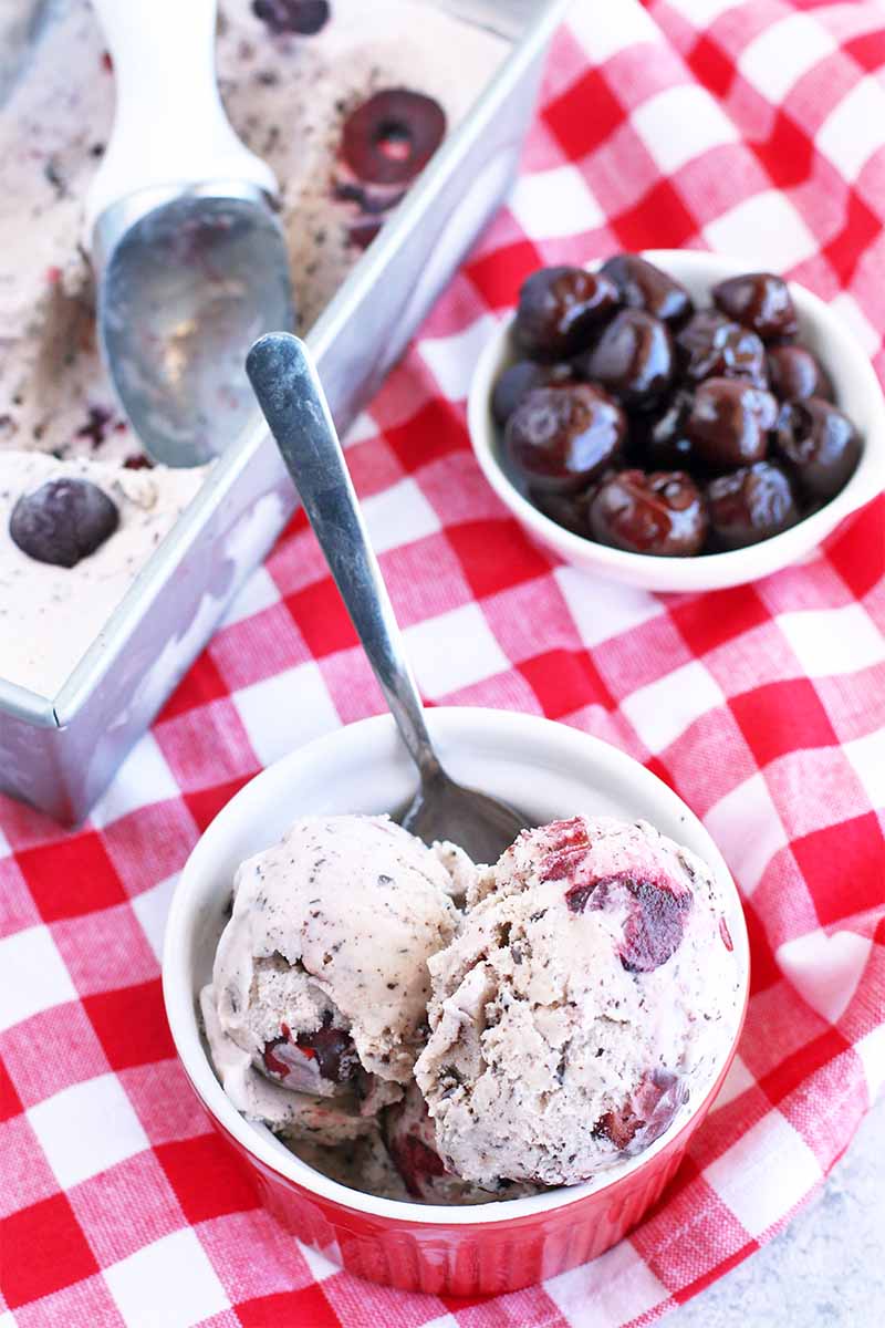 Vertical top-down image of a white bowl of ice cream with a spoon stuck into it with a small white bowl of cherries and more of the frozen dessert with a scoop in a metal loaf pan in the background, on a red and white checkered tablecloth.