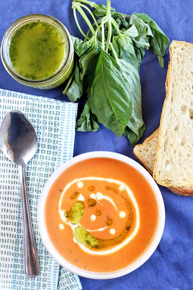 Top-down shot of a bowl of tomato bisque garnished with cream and basil oil, with a spoon on a folded gray and white cloth napkin, a jar of green herb oil, sprigs of fresh basil, and slices of sourdough bread, on a blue cloth background.