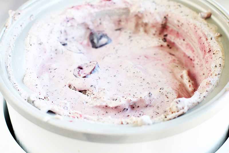 A pale pink ice cream mixture in a frozen white and gray canister, with pieces of cherry and chocolate.