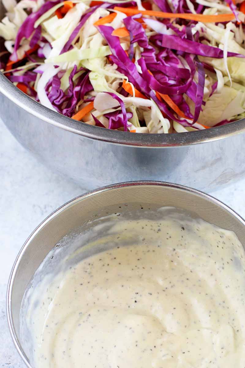 Vertical oblique image of a large stainless steel bowl of shredded cabbage and carrots, and a smaller bowl of mayonnaise dressing.