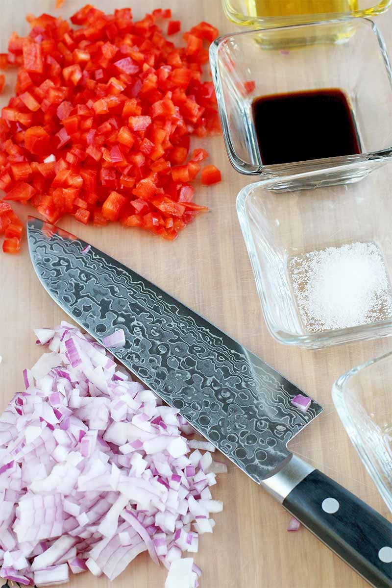 A chef's knife with diced red onion and bell pepper, on a beige countertop with small square glass dishes of balsamic vinegar and salt.