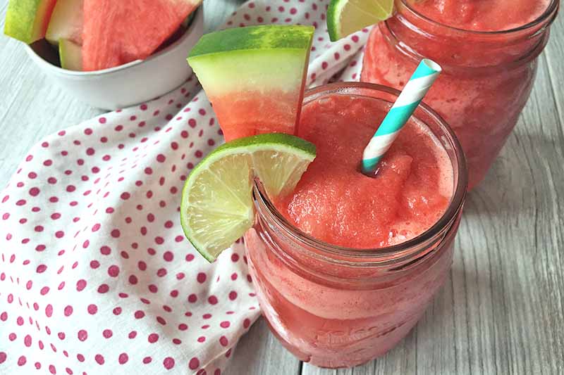 Horizontal image of watermelon daiquiris in glass jars with garnishes on a gray wooden surface with a spotted napkin.