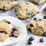 Blueberry scones on a white plate, a metal cooling rack, and a white piece of parchment paper, with a few scattered raw berries in the foreground.