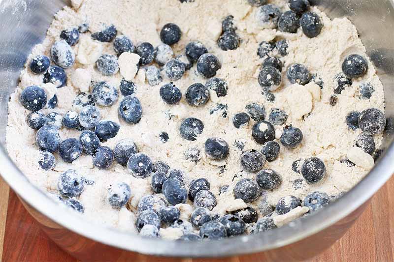 Blueberries coated with flour and stirred into a dry flour and butter mixture in a large stainless steel mixing bowl, on a brown wood surface.