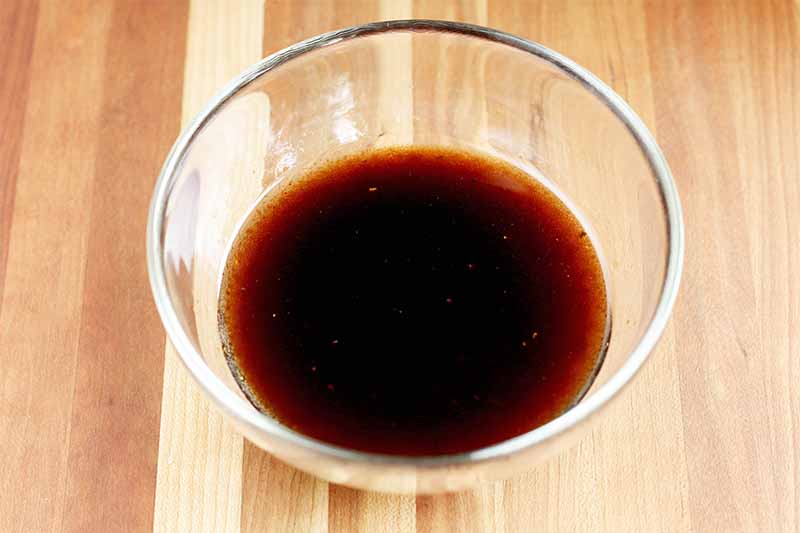 A small glass bowl of balsamic vinegar and olive oil salad dressing, on a striped wood background.