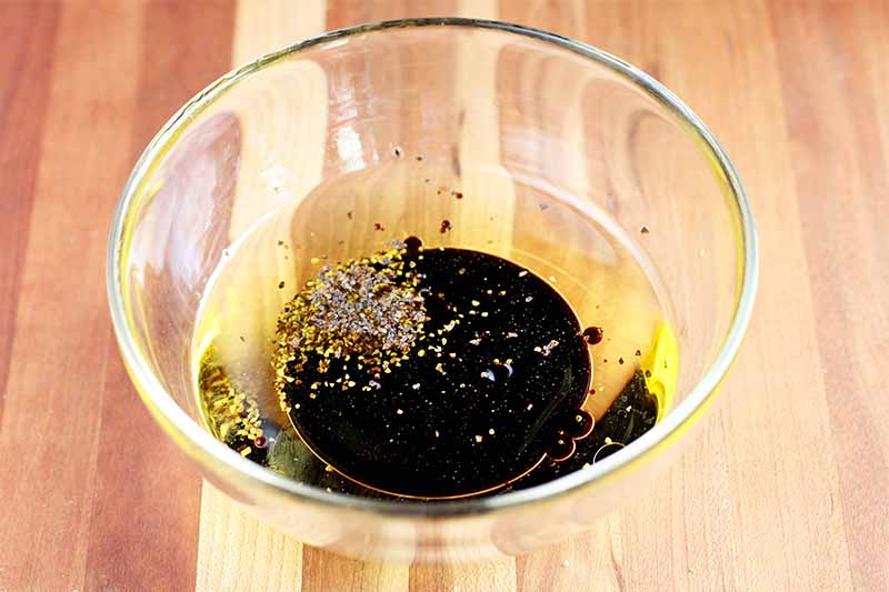 A small glass bowl of olive oil, balsamic vinegar, and black pepper, on a wood background.