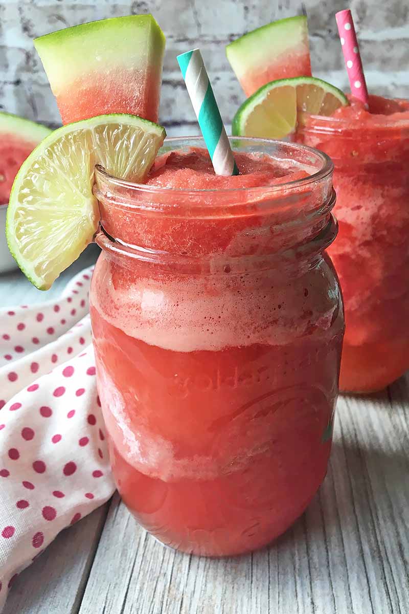 Vertical image of a glass jar filled with watermelon daiquiri with a lime and watermelon wedge.
