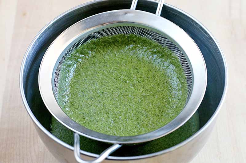 A green basil mixture straining into a stainless steel bowl through a stainless mesh strainer, on a beige countertop.