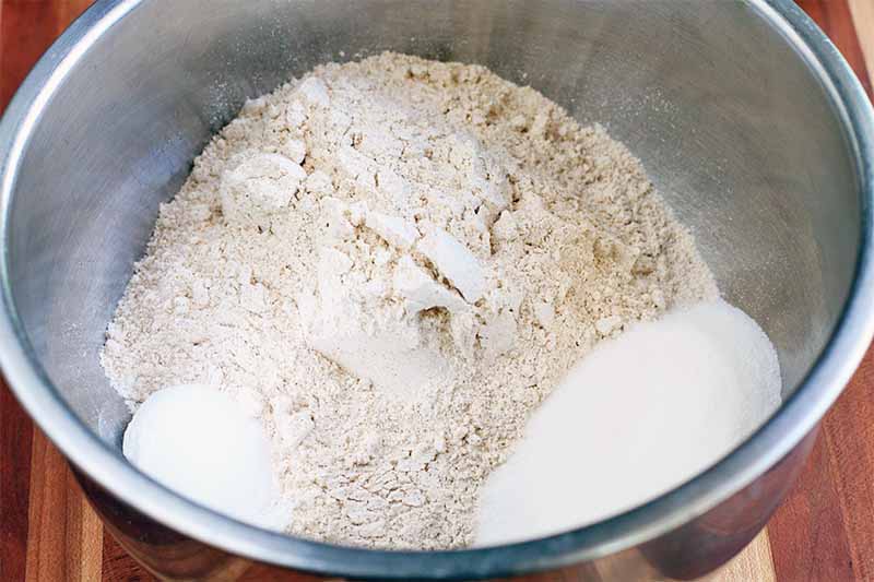 A crumbly flour and butter mixture in a large stainless steel mixing bowl with small piles of sugar and salt, on a brown wood surface.