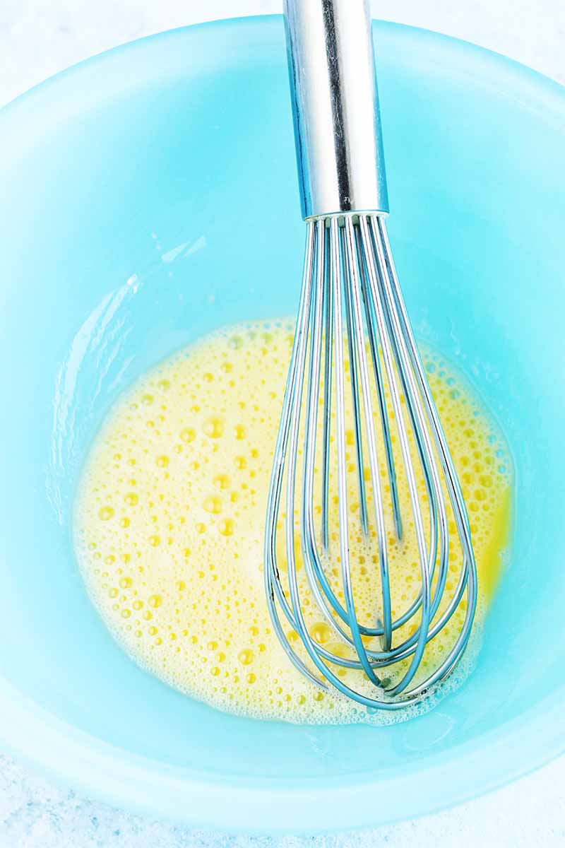 Vertical image of yellow beaten egg in a light blue glass bowl with a wire whisk.