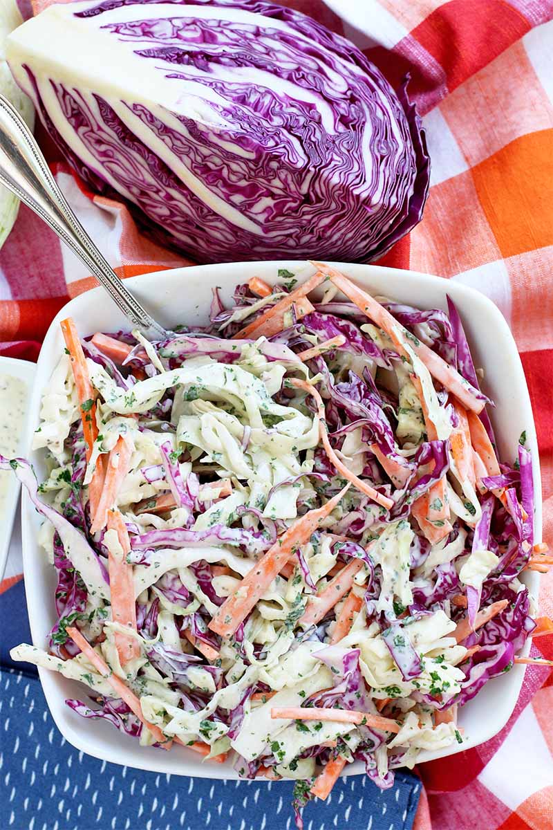 Vertical top-down image of a square dish with rounded corners, filled with homemade cabbage slaw with a mayonnaise sauce, on a red and white checkered cloth background with a quarter of a head of purple cabbage at the top of the frame and a folded blue cloth napkin with white polka dots at the bottom.