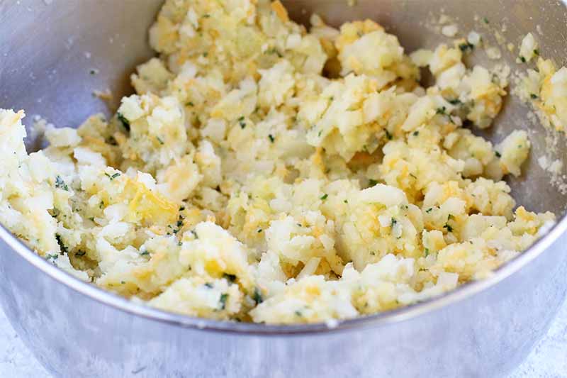 A mashed potato mixture with cheese and herbs in a stainless steel mixing bowl.