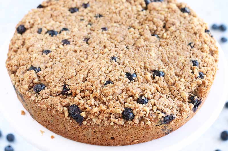 A round blueberry coffee cake on a white cake stand, with fresh berries in shallow focus.
