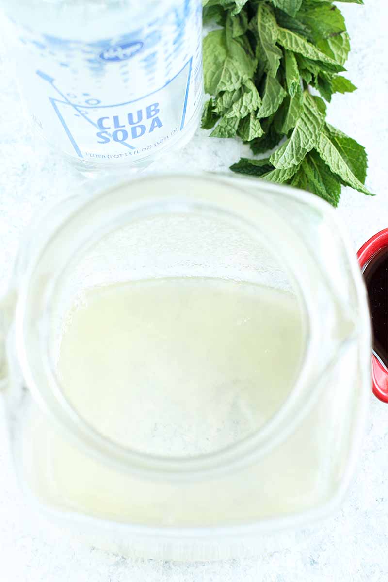 Top-down image of a glass pitcher of a pale yellow liquid next to a clear plastic bottle of club soda and a bunch of fresh mint.