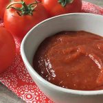 Horizontal image of ketchup in a white bowl on a red napkin with fresh tomatoes.