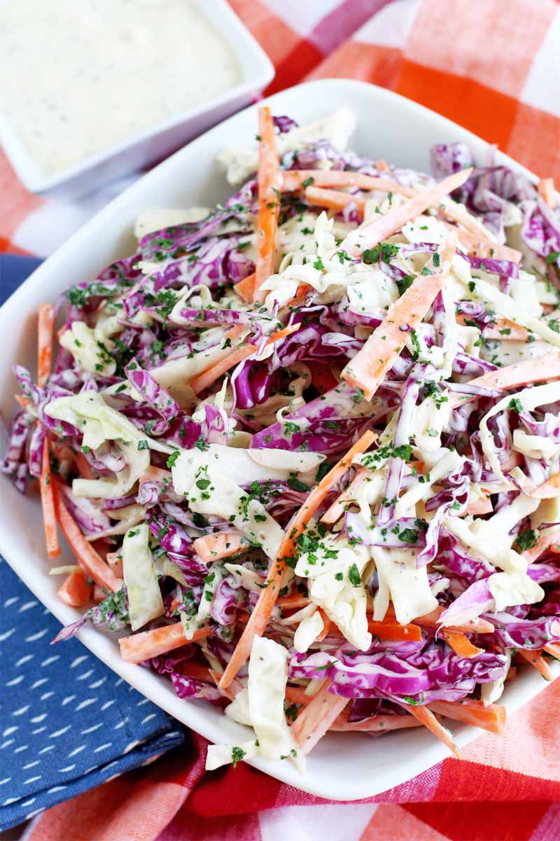 Vertical top down image of red and green cabbage and orange carrot coleslaw in a white square bowl, on a red checkered tablecloth with a folded blue cloth napkin and a small square dish of mayonnaise dressing.